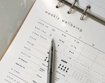 May include: A weekly wellbeing planner with a clear pen on the page. The planner includes sections for wake up, bed time, sleep hours, mood, energy, water, exercise, vitamins, meditation, and a treat. The text is in black.