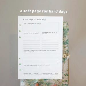 Puede incluir: Una página de planificador en papel blanco con el texto "a soft page for hard days" y indicaciones para la reflexión. La página está abierta, revelando una cubierta con estampado floral con cierre de botón. La página incluye indicaciones para la reflexión.