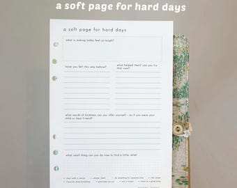 May include: A white paper planner page with the text "a soft page for hard days" and prompts for reflection. The page is held open, revealing a floral patterned cover with a button closure. The page includes prompts for self-reflection.