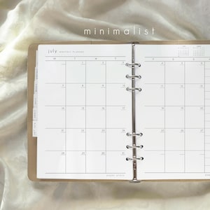 May include: Open planner with a July monthly calendar page. The planner has a minimalist design with a light brown cover and a metal binder. The text "minimalist" is at the top. The calendar grid is black on white.