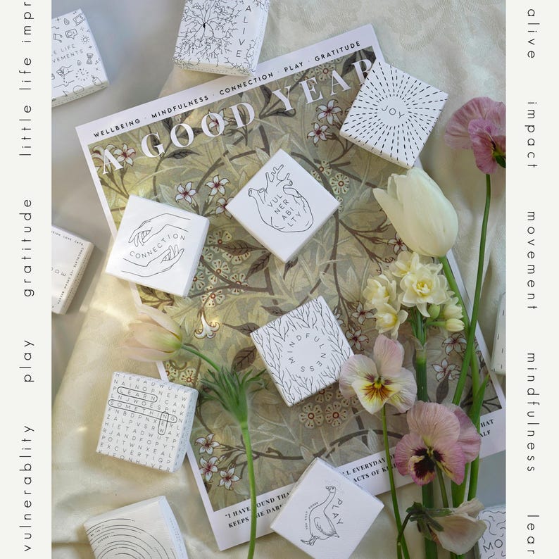 May include: An overhead shot features several small, white square boxes with black line drawings and text, arranged on a floral patterned background. The boxes have words like "Connection" and "Joy". Flowers and the words "A Good Year" are also visible.