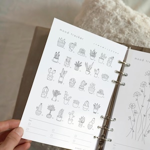 May include: A white planner insert with a "mood tracker" design. The page features numerous small, black-and-white illustrations of cacti and succulents. The planner is open, revealing a second page with floral line art.