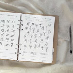 May include: Open planner with a 30-day challenge to plant a forest, featuring tree illustrations. The planner is open to a page with a nature theme, including drawings of insects and plants. A pen rests on the right side.