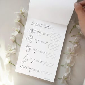 Peut inclure: Un bloc-notes blanc avec le texte "5 senses mindfulness" et des illustrations des cinq sens. Le bloc-notes est ouvert, avec des lignes pour écrire. Des fleurs blanches sont disposées autour du bloc-notes.