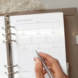 May include: A goal planner page inside a ring binder with a metal mechanism. The page features a header labeled goal planner, a large goal box, three stage boxes, and three task lists with checkboxes. A silver pen is positioned over the paper.
