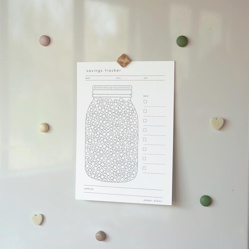 May include: A white savings tracker sheet with a jar illustration, held by magnets on a white surface. The jar is filled with circles, and the sheet has sections for goals and a key. The text "savings tracker" and "paper plans" are visible.