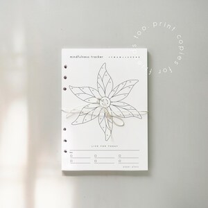 Puede incluir: Un rastreador de atenci&oacute;n plena blanco con un dise&ntilde;o floral, atado con una cuerda. El rastreador incluye el texto "mindfulness tracker" y "LIVE FOR TODAY". El dise&ntilde;o presenta una flor con p&eacute;talos numerados y espacio para notas.