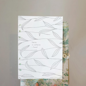 Puede incluir: Una cubierta de planificador blanca con un diseño de hojas negras y el texto "here is where i heal". El planificador está abierto, revelando un interior estampado. Las palabras "paper plans" están en la parte inferior de la cubierta.