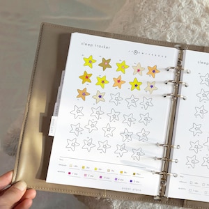 May include: A planner open to a sleep tracker page, with colourful star illustrations. The page is labelled "sleep tracker" and includes a key for tracking sleep quality. The planner is in a beige leather binder.