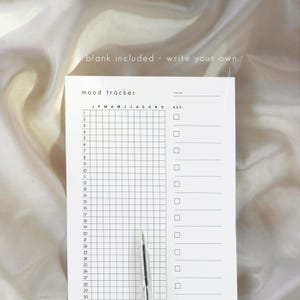 May include: A white mood tracker notepad with a grid for daily entries and a key section. The notepad includes the text "mood tracker" and "blank included - write your own". A pen is positioned near the bottom of the notepad.