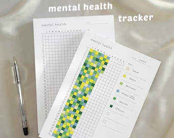 May include: Two white mental health tracker sheets with grid layouts and the text "mental health." One sheet has a color-coded key for tracking moods. A silver pen rests on the side. The word "tracker" is at the top.