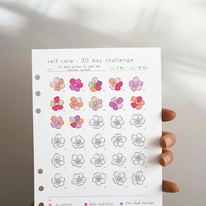 May include: A white paper planner insert titled self care - 30 day challenge with a goal of 2+ daily actions to calm my nervous system. It features a grid of thirty flower outlines, some colored in pink, orange, and purple, with a key at the bottom.