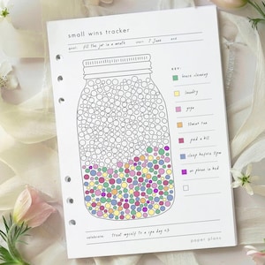 Op de afbeelding: Een witte papieren planner met een "small wins tracker" ontwerp. De tracker heeft een potillustratie gevuld met kleurrijke cirkels, met een sleutel voor verschillende activiteiten. Tekst bevat "fill the jar in a month" en "Treat myself to a spa day <3."