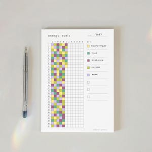 May include: A 2027 energy levels tracker notepad with a grid for daily logging, featuring a colour-coded key for majorly fatigued, tired, mixed energy, energised, and manic states. A clear plastic ballpoint pen is positioned to the left of the pad.