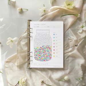 May include: A clear binder holds a savings tracker sheet with a jar graphic filled with colorful dots. The sheet includes a key for different amounts and the text "savings tracker". White flowers and a cream-colored fabric are in the background.