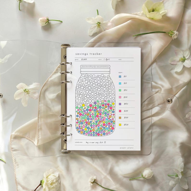 May include: A clear binder holds a savings tracker sheet with a jar graphic filled with colorful dots. The sheet includes a key for different amounts and the text "savings tracker". White flowers and a cream-colored fabric are in the background.