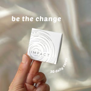 以下が含まれることがあります： 「be the change」と「30 daily cards」の文字が入った、白い「IMPACT」カードの箱。箱には線で描かれた円形のデザインがあります。背景は柔らかいニュートラルカラーです。