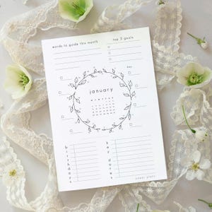 May include: A white paper monthly planner page for January featuring a central circular leaf wreath surrounding the calendar dates. The layout includes sections for words to guide this month, top 3 goals, birthdays, and key dates with checkboxes.