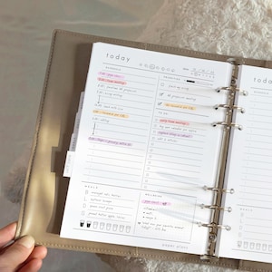 May include: Open planner with a daily schedule, priorities, and to-do list. The planner includes sections for meals and wellbeing, with highlighted entries and the text "paper plans."