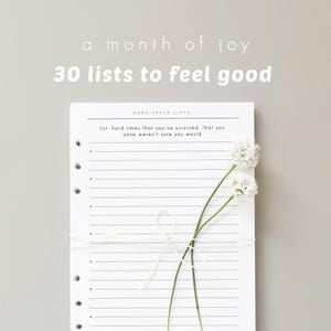 May include: A white paper list pad with punched holes on the left side, featuring the text 'a month of joy', '30 lists to feel good', and 'GOOD FEELS LISTS'. The page includes a prompt about surviving hard times and is tied with white string.