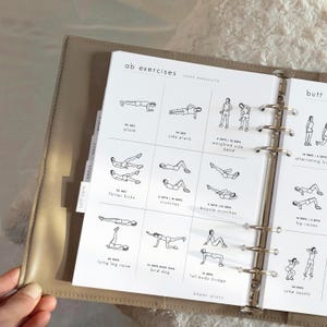 Puede incluir: Un planificador beige con una página de ejercicios abdominales. La página presenta dibujos lineales negros de ejercicios como planchas, abdominales y elevaciones de piernas. El planificador tiene pestañas etiquetadas para el cuidado personal, objetivos y tareas diarias.