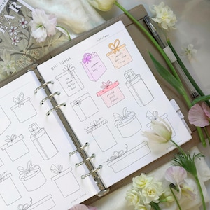 May include: A white planner page featuring black ink line drawings of various gift boxes with bows. The page includes the text "gift ideas" and "christmas" with some boxes filled in with pink and orange colours and handwritten text labels.