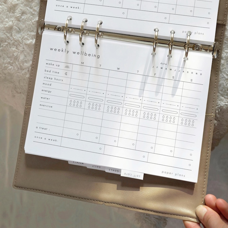 May include: A beige planner with a weekly wellbeing insert. The insert has a grid layout for tracking daily habits, including wake up time, sleep hours, mood, energy, water intake, and exercise. The planner includes tabs labeled "Self Care", "Goals", and "Daily".