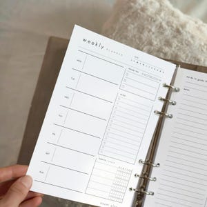May include: A white weekly planner page with black text, open in a binder. The planner includes sections for each day of the week, priorities, to-do lists, and habit tracking. The words "weekly planner" are at the top.