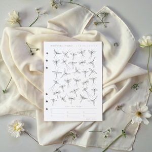 Op de afbeelding: Een witte mindfulness tracker met een illustratie van een libel. De tracker heeft de tekst "mindfulness tracker" en "Live for Today". Het ligt op een crèmekleurige doek met witte bloemen.