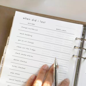 May include: A planner page with the text "when did I last" and a list of actions to track, including "change toothbrush" and "wash bedding." The page is open in a binder, with a pen resting on the page. The planner has tabs labeled "Daily," "Goals," and "Weekly."