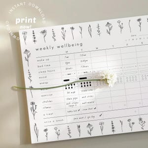 May include: A white weekly wellbeing planner with black floral illustrations and text. The planner includes sections for wake up, bed time, sleep hours, mood, energy, exercise, stretches, vitamins, and a treat. A white flower and stem are placed on the planner.