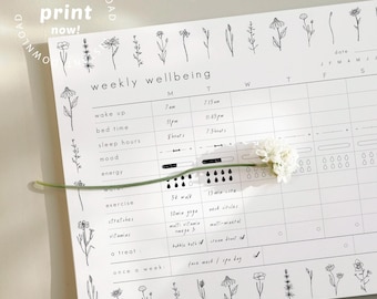 May include: A white weekly wellbeing planner with black floral illustrations and text. The planner includes sections for wake up, bed time, sleep hours, mood, energy, exercise, stretches, vitamins, and a treat. A white flower and stem are placed on the planner.