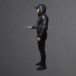 May include: A black leather hooded jacket with matching black pants and a black baseball cap. The jacket has a zipper closure and the hood is up. The person is wearing black sneakers.