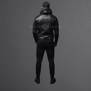May include: A black leather hoodie and matching black pants. The hoodie has a hood and a zipper closure. The pants are fitted. The person is wearing a black baseball cap. The text "Italy Made Leather" is visible.