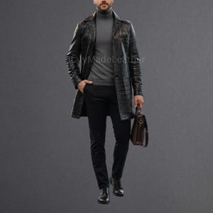Könnte beinhalten: Ein schwarzer Ledermantel mit Krokodiltextur. Der Mantel wird über einem grauen Rollkragenpullover und einer schwarzen Hose getragen. Eine braune Lederaktentasche wird in der rechten Hand gehalten. Das Bild ist vor grauem Hintergrund.