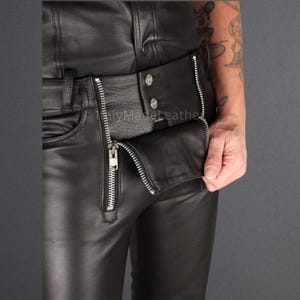 May include: Black leather pants with a silver zipper and button closure. The pants have a classic five-pocket design and a fitted silhouette. The zipper is partially unzipped, revealing the interior of the pants.