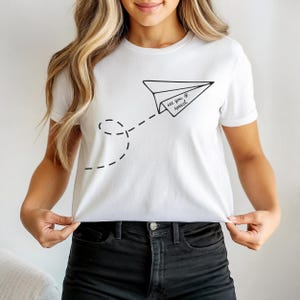 Peut inclure: T-shirt blanc avec un motif d'avion en papier noir et le texte "see you speech". L'avion a une ligne pointillée qui le suit. Le t-shirt est de style col rond classique.