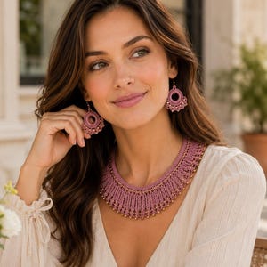 May include: A mauve crochet necklace and matching circular drop earrings. The necklace features a wide, textured collar design with small gold-toned beads integrated along the lower edge. The earrings mirror the circular motif of the necklace.