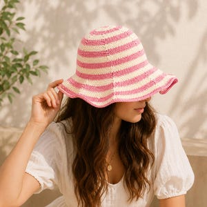 Puede incluir: Un sombrero de pescador tejido a ganchillo con rayas horizontales en tonos alternos de rosa suave y crema. El sombrero presenta una copa redondeada y un ala ancha ligeramente ondulada. La textura del tejido es visible en toda la pieza.