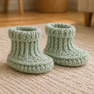 Può includere: Due scarpine da neonato lavorate a maglia verde salvia. Le scarpine sono realizzate con un filato morbido e hanno un design a coste. Le scarpine sono su un tappeto beige. Queste scarpine sono un accessorio accogliente e confortevole per un bambino.
