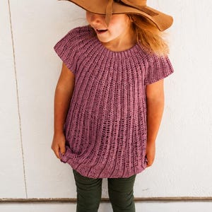 May include: A mauve short-sleeved knitted top featuring a vertical ribbed texture and a round neckline, paired with a wide-brimmed tan felt hat that includes a matching ribbon band tied in a knot at the front, worn over dark green trousers.