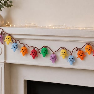 May include: A colorful crochet garland of smiling light bulbs in various colors, including purple, yellow, blue, orange, red, and green. The garland is strung on a brown cord and is displayed on a white mantelpiece.