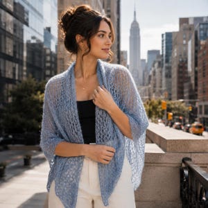 Könnte beinhalten: Eine hellblaue, vorne offene Strickjacke mit einer zarten, luftigen Strickstruktur. Das Kleidungsstück hat lange Ärmel und eine entspannte, drapierte Silhouette, die locker über die Schultern und den Oberkörper fällt und ein feines Garn zeigt.