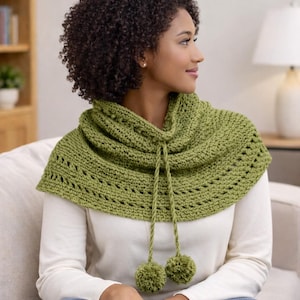 Puede incluir: Un cuello de ganchillo verde con un cordón y dos pompones. El cuello es de color verde claro y tiene un patrón texturizado con agujeros decorativos. El cuello se lleva sobre una camisa blanca de manga larga.