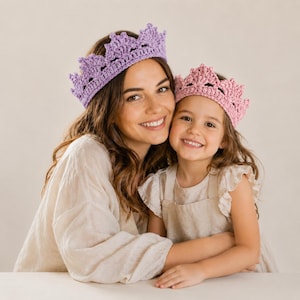 Op de afbeelding: Een vrouw en een kind dragen gehaakte kronen, één lavendelkleurig en één roze. De kronen hebben een open ontwerp. De vrouw draagt een lichtgekleurde blouse en het kind draagt een lichtgekleurde jurk.