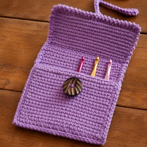 May include: A lavender crochet case featuring a fold-over flap and a dark brown circular button closure. The interior pocket contains three metallic crochet hooks in pink, gold, and silver, with a matching lavender crochet strap attached at the top.