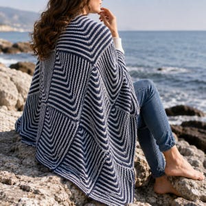 Può includere: Un poncho in maglia a righe blu navy e bianco con un motivo geometrico a chevron, indossato sopra un paio di jeans blu. Il capo presenta un aspetto testurizzato e a coste con un design angolare distinto che ricade sulle spalle e sulla schiena.