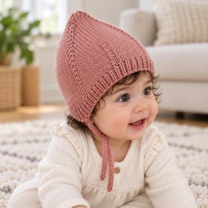 May include: A dusty rose knitted bonnet featuring a pointed crown, ribbed trim along the face opening, and two thin chin ties. The bonnet is crafted with a textured knit pattern and vertical decorative stitching that runs from the top to the base.