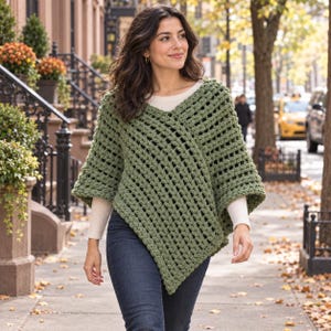 May include: An olive green crochet poncho with a V-neckline and an open-weave pattern, worn over a long-sleeved cream-colored shirt and dark blue denim jeans. The garment features a textured, chunky yarn construction with a pointed hemline.
