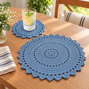 May include: Two round, dusty blue crochet placemats featuring a detailed, intricate circular pattern with a scalloped, pointed edge. The larger placemat is positioned in the foreground, while the smaller one sits behind it, supporting a clear glass of water.